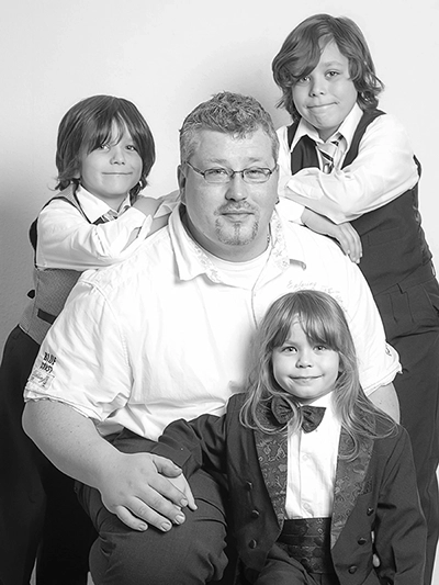 Ein professionell aufgenommenes Studio-Porträt in Schwarz-Weiß, das einen lächelnden Vater mit seinen drei Jungen in Anzügen darstellt, inszeniert von Fotograf Michael Schalansky.